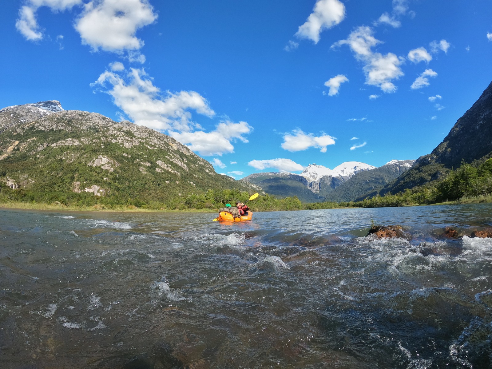 Packraft naranjo remando por las aguas verdes del humedal, con cordillera al fondo